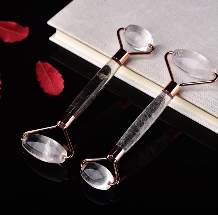 Two crystal quartz facial rollers with rose gold frames, displayed on a black surface with red petals, evoking a sense of elegance and luxury skincare.