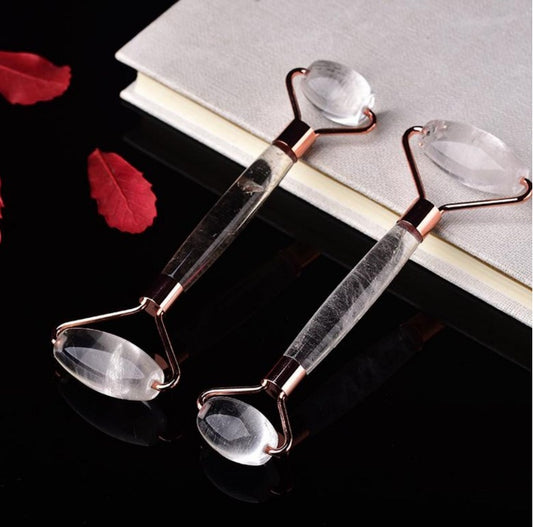 Two crystal quartz facial rollers with rose gold frames, displayed on a black surface with red petals, evoking a sense of elegance and luxury skincare.