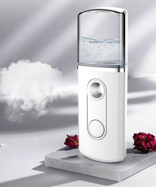 "White portable nano mist facial sprayer releasing fine mist, placed on a marble surface with soft lighting and decorative flowers"