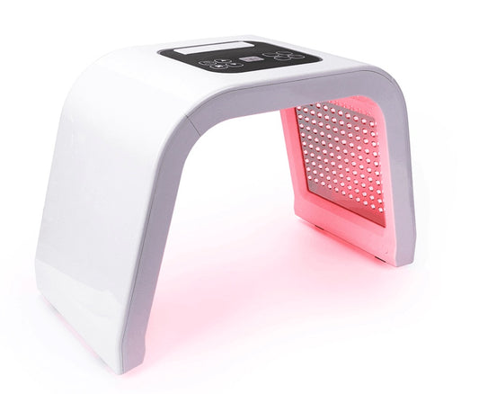 White LED light therapy mask positioned on table with soft pink glow on side panel. Shows detachable design and modern shape, suitable for home skincare routines.